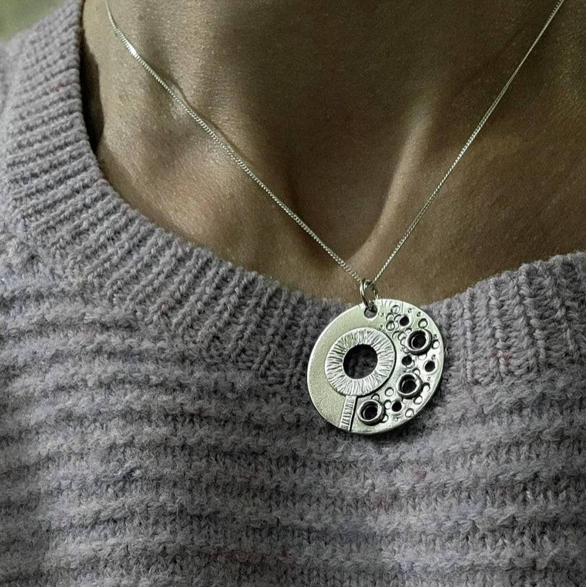 Close-up of a circle silver Necklace worn around a woman's neck atop a sweater.