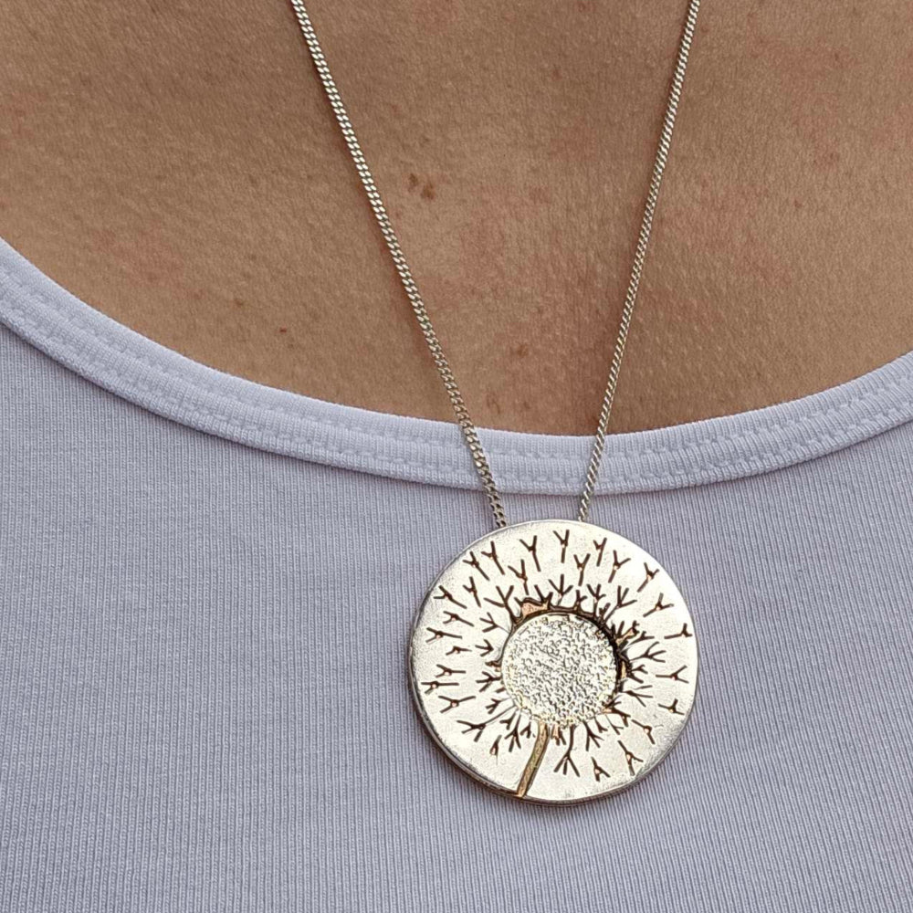 One of a kind handcrafted dandelion necklace in Sterling Silver and Brass on a woman's neck atop a white shirt.