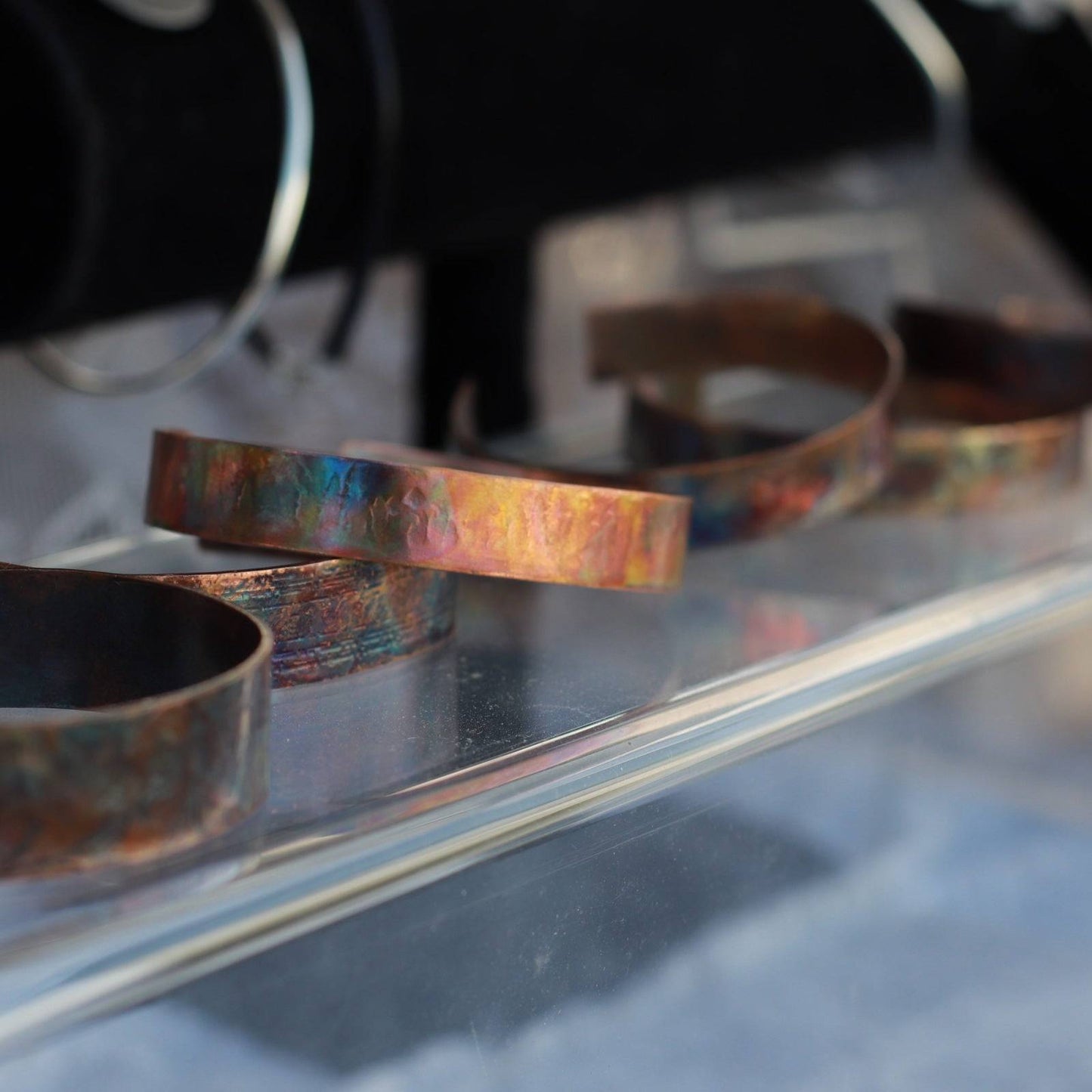 flame painted copper cuff bracelets on acrylic display with another jewelry display blurred behind it.