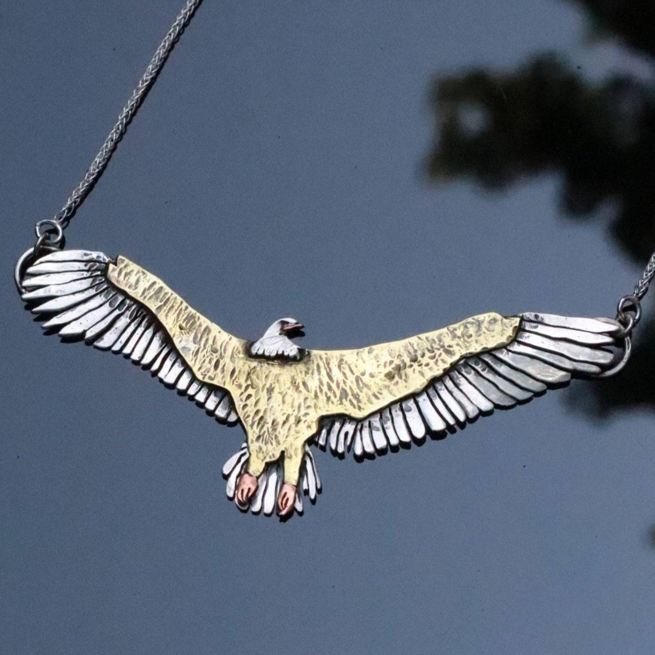 close up of custom jewelry design eagle necklace laid on a reflective board with branches of a tree in the reflection