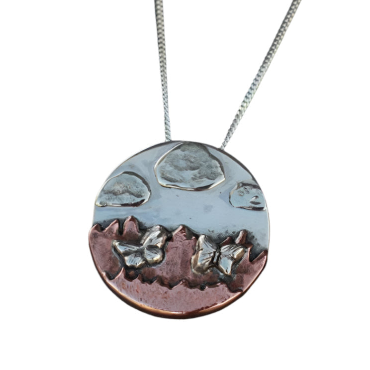 Necklace in silver and copper of a mountainscape with butterflies and clouds.