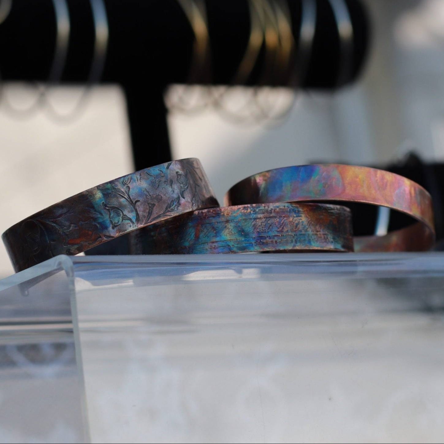 flame painted copper cuff bracelets on acrylic display with another bracelet display blurred in the background.