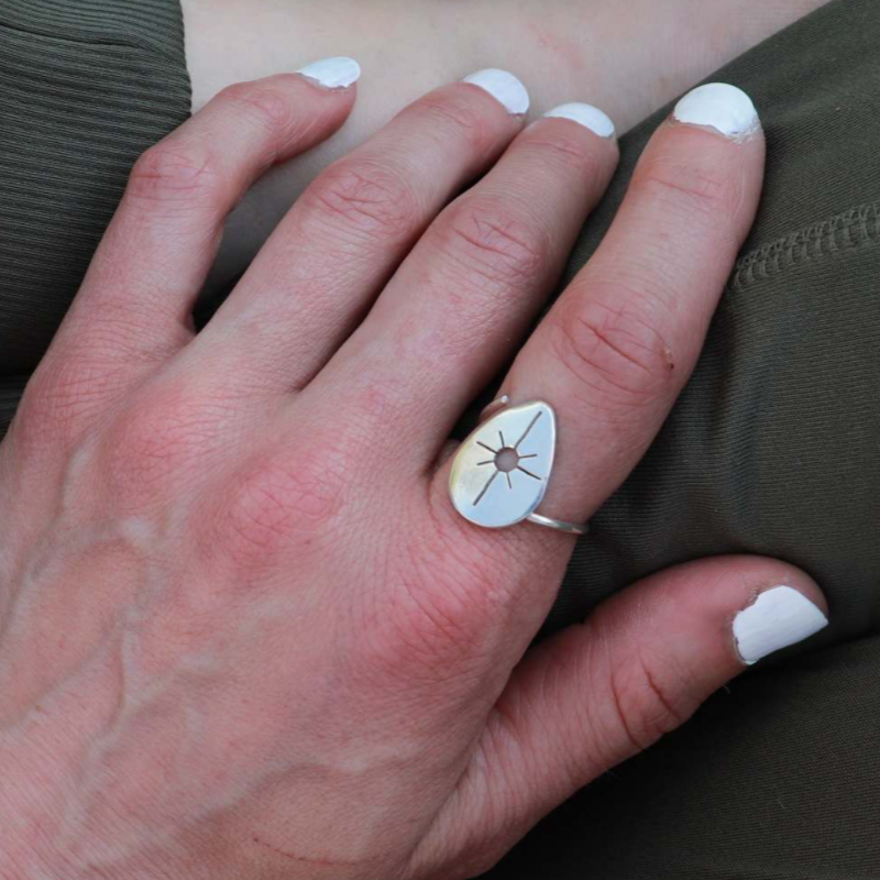 Silver drop ring worn on the pointer finger of a woman resting her hand on crossed legs.